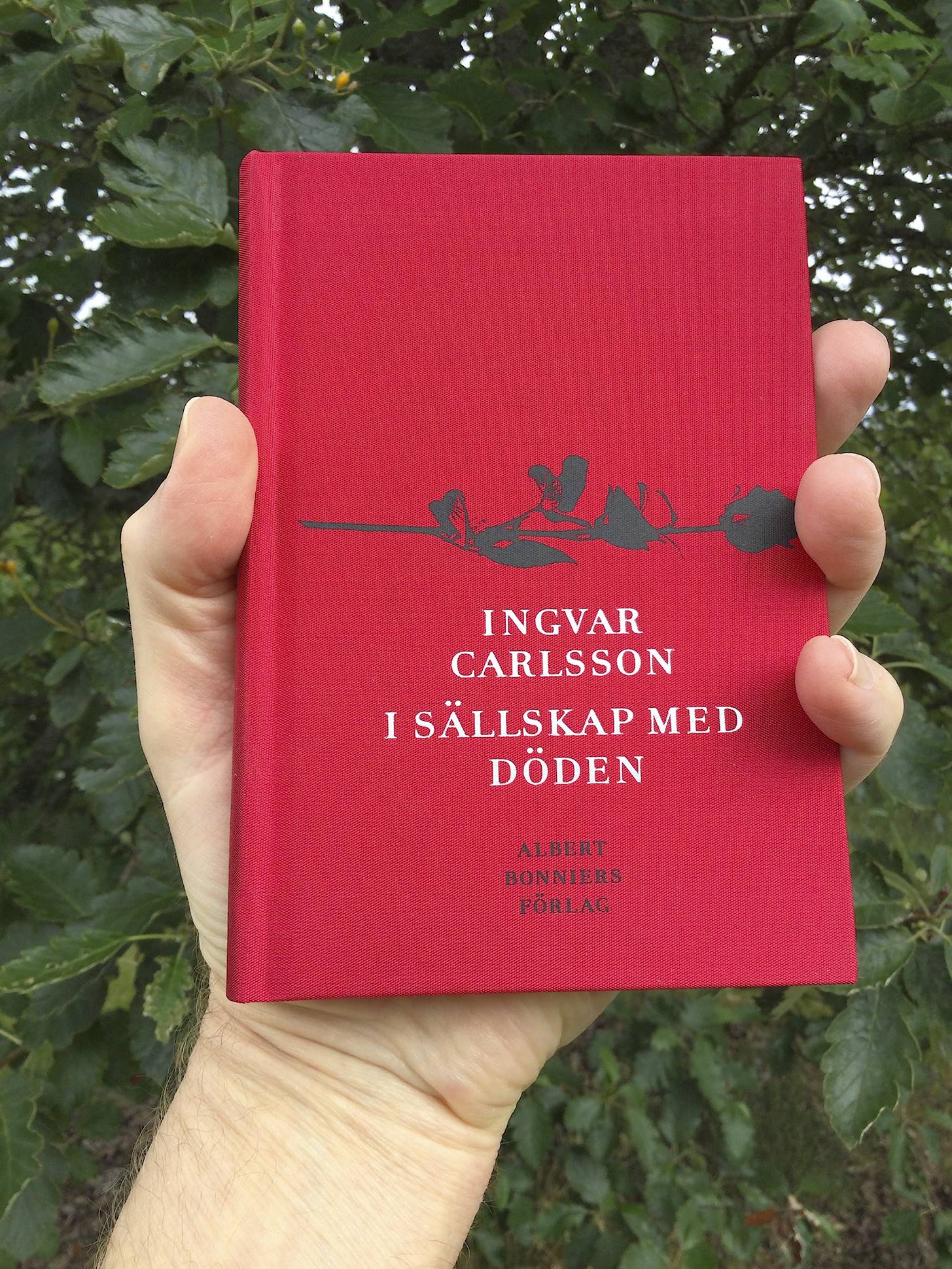 Jag håller den rödfärgade boken I sällskap med döden av Ingvar Carlsson i min hand. Mot en bakgrund av lövverket på ett träd. 